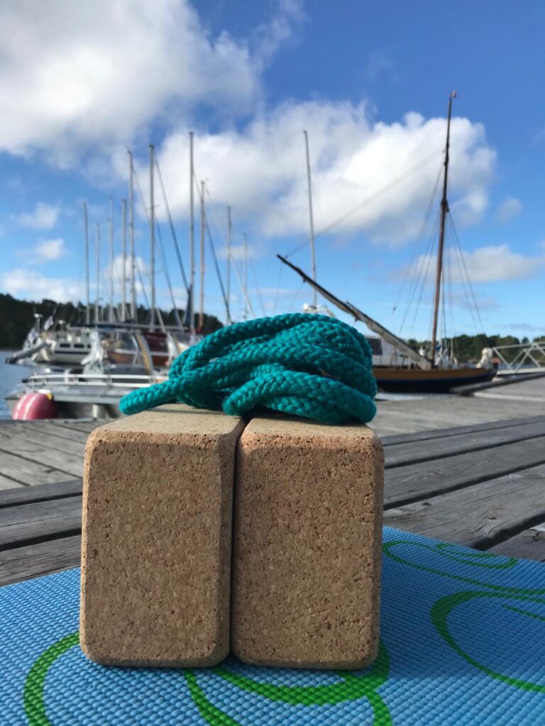 Two cork yoga blocks and a teal rope on a dock, with sailboats and blue sky with clouds in the background.