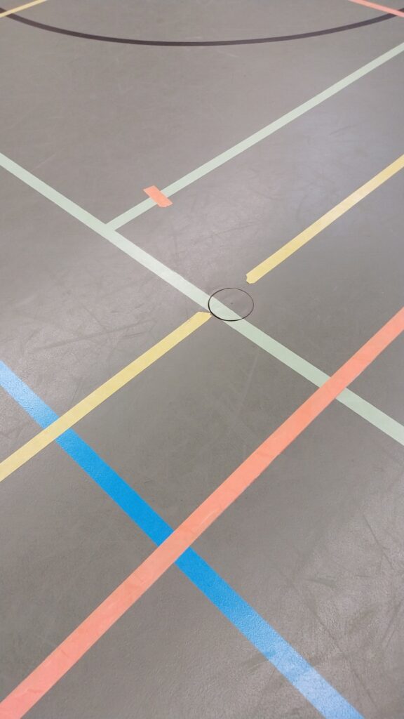 A gym floor with intersecting colored lines in blue, yellow, pink, and green. A round sports equipment socket is visible at the center.