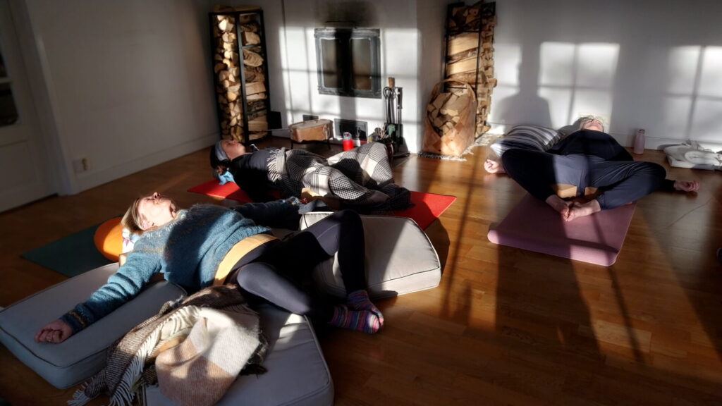 Three people are lying on yoga mats in a sunlit room, relaxing in a restorative yoga pose with props and blankets around them.