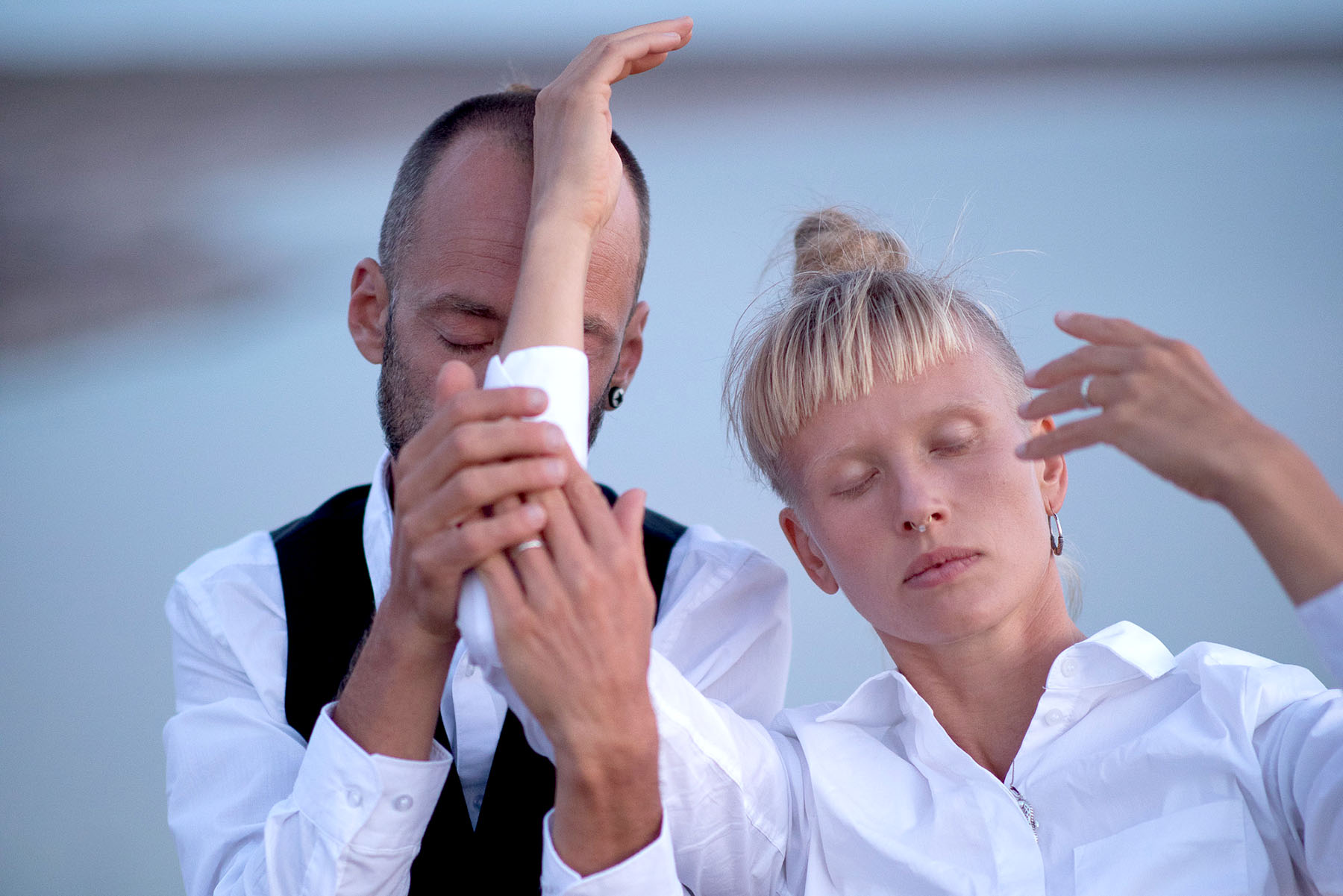 Two people in white shirts with closed eyes pose outdoors, their arms raised and hands touching in an expressive gesture.