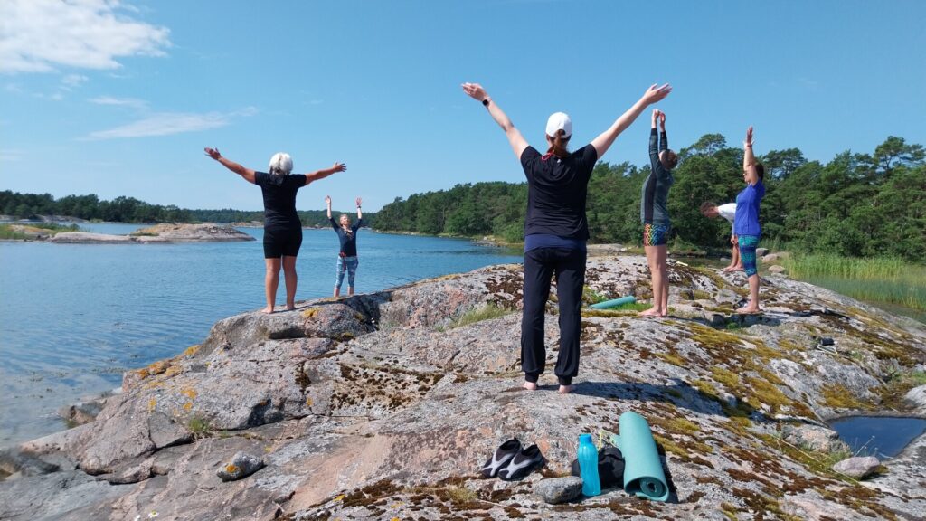 Människor utövar yoga på en klippig strand vid vattnet, med armarna lyfta, under en klarblå himmel. Yogamattor och skor syns i närheten.