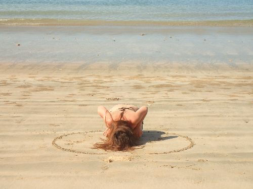 Person lying face down on sandy beach within a drawn circle, near the water's edge.