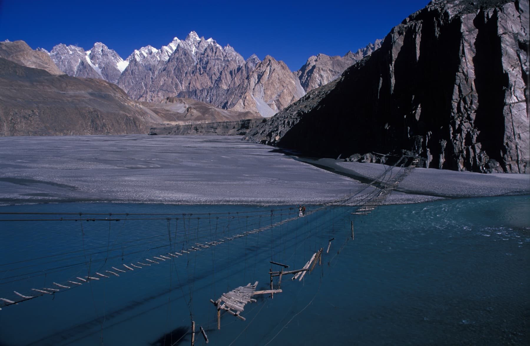 A narrow, rustic suspension bridge crosses a wide river with a mountainous landscape in the background under a clear blue sky.