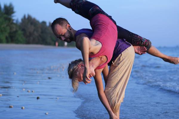 Two people are performing an acrobatic yoga pose on a beach near the water’s edge, with trees and the sea visible in the background.